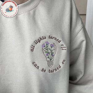 Puede incluir: Una sudadera beige claro con un diseño bordado. El diseño presenta una bombilla con una mano sosteniendo flores, rodeada por el texto "all lights turned off can be turned on". La sudadera tiene cuello redondo.