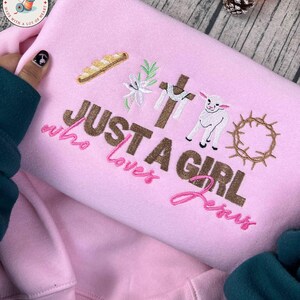 Op de afbeelding: Roze sweatshirt met geborduurde religieuze symbolen, waaronder een kruis, een lam en een doornenkroon. De tekst "JUST A GIRL who loves Jesus" is ook geborduurd. De sweatshirt is gemaakt van een zacht materiaal.
