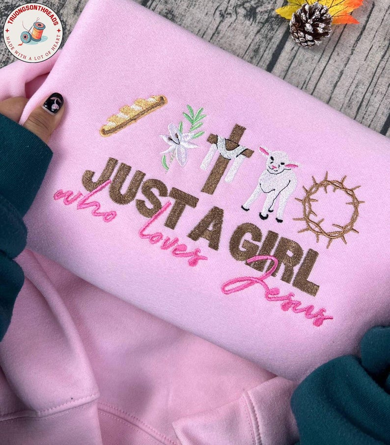 Op de afbeelding: Roze sweatshirt met geborduurde religieuze symbolen, waaronder een kruis, een lam en een doornenkroon. De tekst "JUST A GIRL who loves Jesus" is ook geborduurd. De sweatshirt is gemaakt van een zacht materiaal.