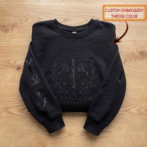 Könnte beinhalten: Ein schwarzes Sweatshirt mit Rundhalsausschnitt und tonaler schwarzer Stickerei. Auf der Brust befindet sich ein Drache in einem kreisförmigen Kranz und der Text BASGIATH WAR COLLEGE. Die Ärmel zeigen gestickte Drachen und einen Dolch mit dem Text Violent Little Things.