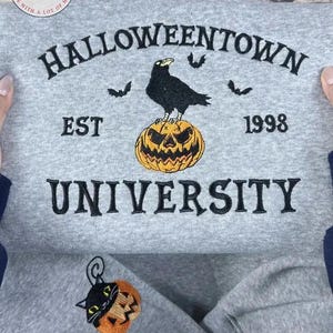 K&ouml;nnte beinhalten: Ein graues Sweatshirt mit dem gestickten Text "Halloweentown University EST 1998". Das Design zeigt eine schwarze Kr&auml;he auf einem orangefarbenen K&uuml;rbis und drei Flederm&auml;use. Ein kleiner gestickter Aufn&auml;her mit einer schwarzen Katze in einem K&uuml;rbis ist ebenfalls sichtbar.
