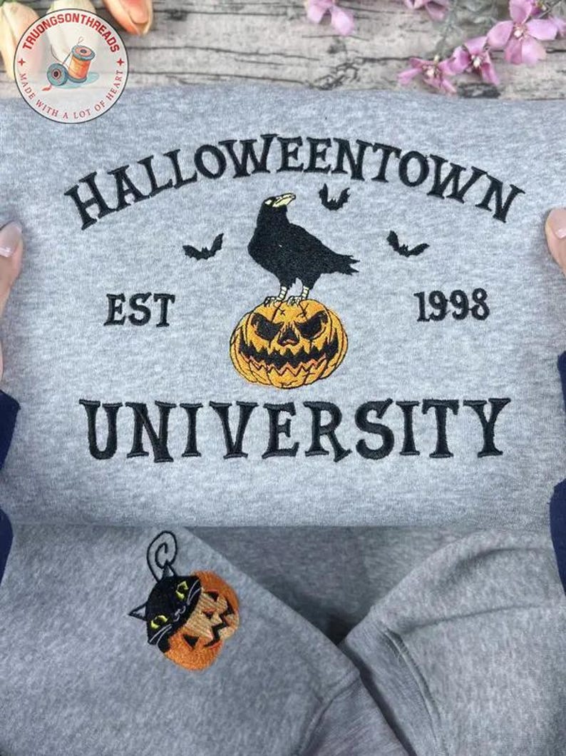 K&ouml;nnte beinhalten: Ein graues Sweatshirt mit dem gestickten Text "Halloweentown University EST 1998". Das Design zeigt eine schwarze Kr&auml;he auf einem orangefarbenen K&uuml;rbis und drei Flederm&auml;use. Ein kleiner gestickter Aufn&auml;her mit einer schwarzen Katze in einem K&uuml;rbis ist ebenfalls sichtbar.