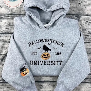 K&ouml;nnte beinhalten: Grauer Kapuzenpullover mit dem Text "Halloweentown University" und einem Kr&auml;hen-auf-K&uuml;rbis-Design. Der linke &Auml;rmel zeigt einen K&uuml;rbis mit einer Katze. Der Hoodie hat eine Kapuze mit Kordelzug.