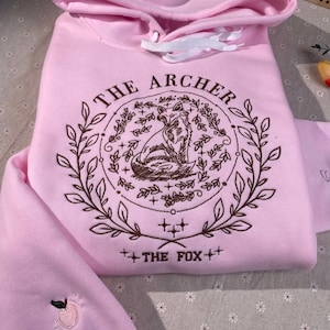 May include: A pink hoodie with a brown embroidered design featuring a fox in a circle of leaves and the text "THE ARCHER" and "THE FOX".
