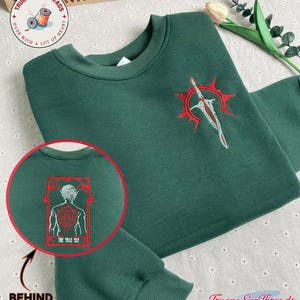 Puede incluir: Una sudadera verde oscuro con diseños bordados en rojo. El diseño frontal presenta una espada y un sol. El diseño trasero presenta una figura con el texto "The Tall Elf".