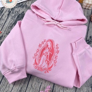 May include: A light pink hoodie with a drawstring hood and long sleeves. The front features a red graphic of a religious figure surrounded by floral elements. The hoodie is made of a soft material and is suitable for casual wear.