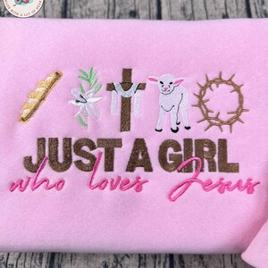 Op de afbeelding: Zachtroze sweatshirt met geborduurde religieuze symbolen: brood, lelie, kruis, lam en doornenkroon. De tekst luidt: "JUST A GIRL who loves Jesus".