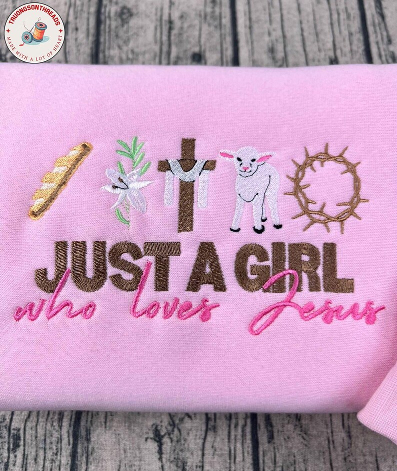 Op de afbeelding: Zachtroze sweatshirt met geborduurde religieuze symbolen: brood, lelie, kruis, lam en doornenkroon. De tekst luidt: "JUST A GIRL who loves Jesus".