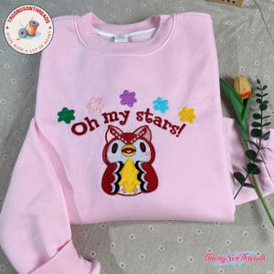 Puede incluir: Sudadera rosa claro con un dise&ntilde;o de b&uacute;ho bordado y la frase "Oh my stars!". El b&uacute;ho es rojo, amarillo y blanco, rodeado de adornos en forma de estrella de colores. Tambi&eacute;n se ve la marca "Truong Son Threads".