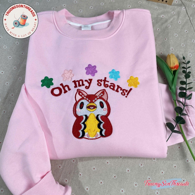 Puede incluir: Sudadera rosa claro con un dise&ntilde;o de b&uacute;ho bordado y la frase "Oh my stars!". El b&uacute;ho es rojo, amarillo y blanco, rodeado de adornos en forma de estrella de colores. Tambi&eacute;n se ve la marca "Truong Son Threads".