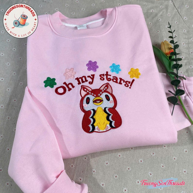 Puede incluir: Una sudadera rosa claro con la frase bordada "Oh my stars!" y un dise&ntilde;o de b&uacute;ho de dibujos animados. El b&uacute;ho es rojo, blanco y amarillo, con acentos de estrellas de colores. La sudadera tiene cuello redondo y mangas largas.