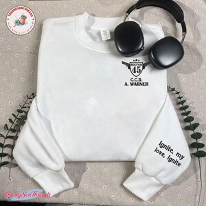 May include: A white sweatshirt with the text "SECTOR 45 C.C.R. A. WARNER" on the chest and "Ignite, my love, Ignite" on the sleeve. Black headphones rest on the sweatshirt. The sweatshirt has a crew neck and long sleeves.