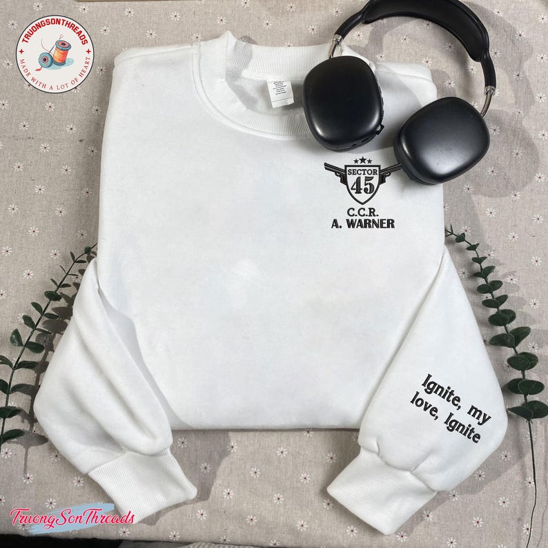May include: A white sweatshirt with the text "SECTOR 45 C.C.R. A. WARNER" on the chest and "Ignite, my love, Ignite" on the sleeve. Black headphones rest on the sweatshirt. The sweatshirt has a crew neck and long sleeves.