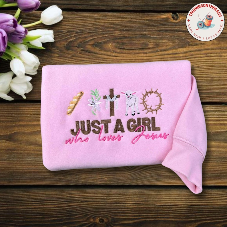 Op de afbeelding: Een roze sweatshirt met geborduurde religieuze symbolen en de tekst "JUST A GIRL who loves Jesus." Het ontwerp omvat een kruis, een lam, een doornenkroon en brood. De sweatshirt ligt op een houten oppervlak.