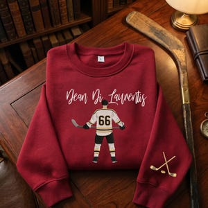 May include: A burgundy sweatshirt with a hockey player graphic and the name "Dean Di Laurentis" in white script. The player wears a white, black, and gold uniform with the number 66. Two crossed hockey sticks are on the sleeve.