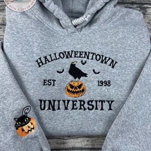 K&ouml;nnte beinhalten: Grauer Hoodie zum Thema Halloween mit dem Text "Halloweentown University" in Schwarz gestickt. Eine schwarze Kr&auml;he sitzt auf einem orangefarbenen K&uuml;rbis. Auf dem &Auml;rmel ist ein kleiner K&uuml;rbis mit einer schwarzen Katze gestickt.