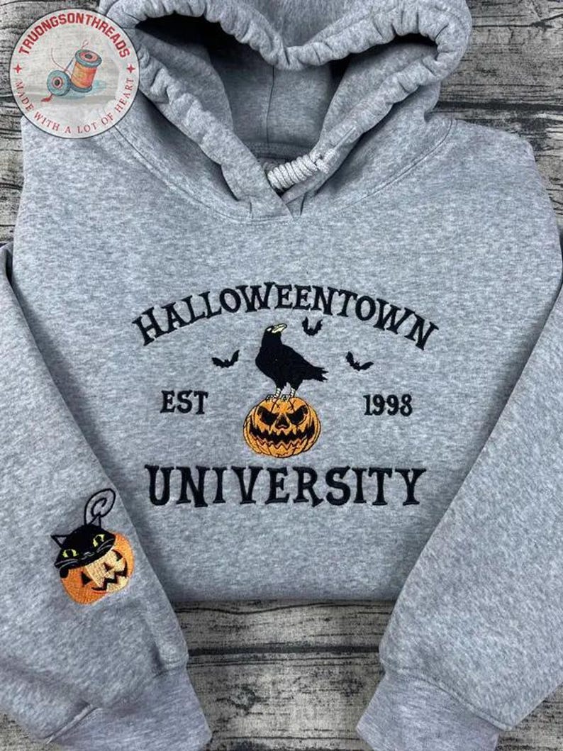 K&ouml;nnte beinhalten: Grauer Hoodie zum Thema Halloween mit dem Text "Halloweentown University" in Schwarz gestickt. Eine schwarze Kr&auml;he sitzt auf einem orangefarbenen K&uuml;rbis. Auf dem &Auml;rmel ist ein kleiner K&uuml;rbis mit einer schwarzen Katze gestickt.