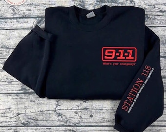 May include: Black sweatshirt with red embroidered text and graphics. The front features "9-1-1" inside a rectangle, with "What's your emergency?" below. The left sleeve reads "STATION 118" with names below.