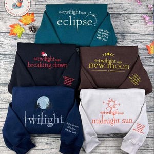 Puede incluir: Sudaderas plegadas en verde azulado, negro, marrón, azul marino y blanco, cada una bordada con texto y diseños de la serie "The Twilight Saga". Cada sudadera muestra el título de un libro, como "Eclipse" o "Breaking Dawn".