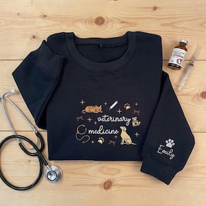 Puede incluir: Una sudadera negra de manga larga con bordados dorados de un gato, un perro, un estetoscopio, una jeringuilla y un frasco de medicina, el texto "veterinary medicine" en el centro y el nombre "Emily" con una huella de pata en la manga.