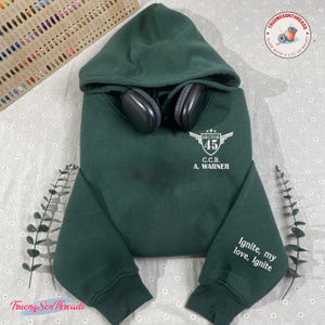 May include: A dark green hooded sweatshirt with the text "SECTOR 45 C.C.R. A. WARNER" and "Ignite, my love, Ignite" printed in white. Black headphones rest on the hood. The sweatshirt is displayed on a patterned surface.