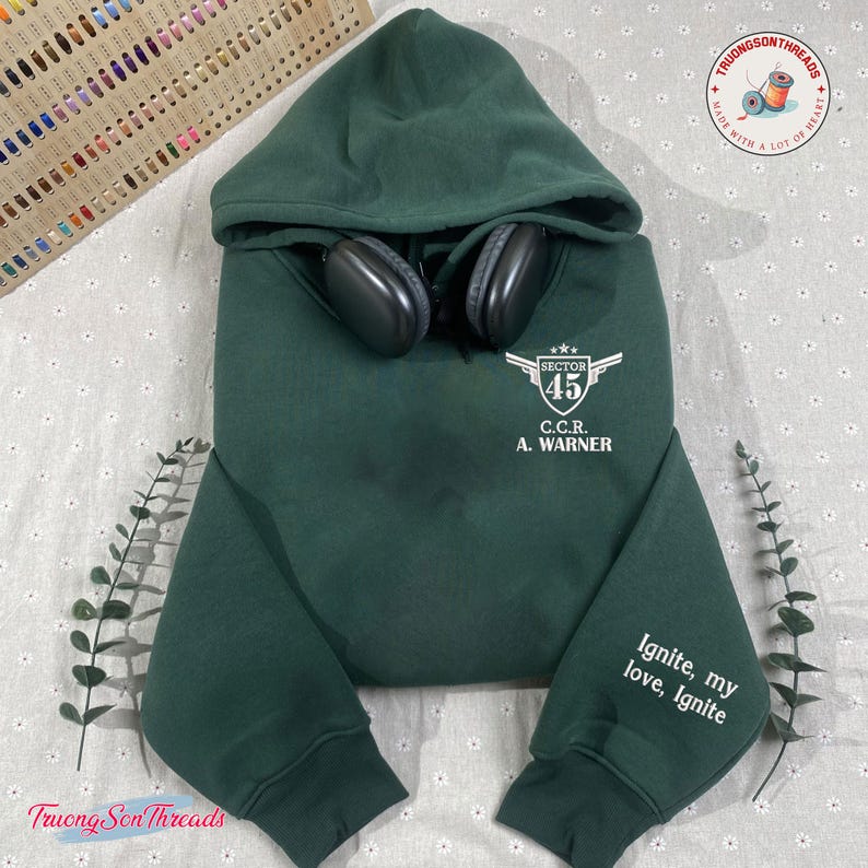 May include: A dark green hooded sweatshirt with the text "SECTOR 45 C.C.R. A. WARNER" and "Ignite, my love, Ignite" printed in white. Black headphones rest on the hood. The sweatshirt is displayed on a patterned surface.