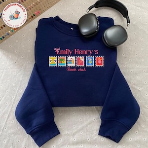 Puede incluir: Sudadera azul marino con el texto "Emily Henry's Book club" en rosa. El diseño incluye ilustraciones de sellos de colores. Auriculares negros descansan sobre la sudadera.