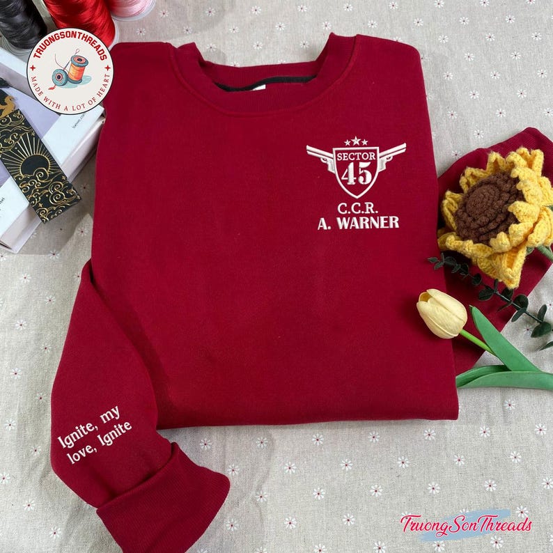 May include: A maroon sweatshirt with white text and graphics. The front features a winged emblem with "SECTOR 45" and "C.C.R. A. WARNER." The left sleeve reads "Ignite, my love, Ignite." The sweatshirt is displayed on a floral patterned surface.
