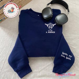 May include: Navy blue sweatshirt with white text that reads "SECTOR 45 C.C.R. A. WARNER" on the chest. The sleeve has the text "Ignite, my love, Ignite". A pair of black headphones rests on the sweatshirt.
