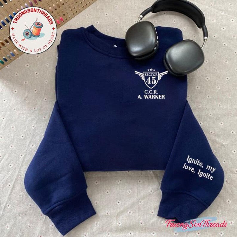 May include: Navy blue sweatshirt with white text that reads "SECTOR 45 C.C.R. A. WARNER" on the chest. The sleeve has the text "Ignite, my love, Ignite". A pair of black headphones rests on the sweatshirt.