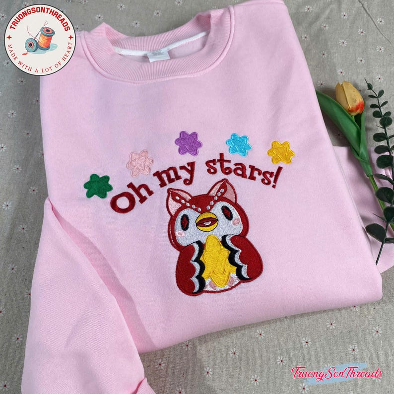 Puede incluir: Una sudadera rosa claro con la frase bordada "Oh my stars!" y un b&uacute;ho de dibujos animados. El b&uacute;ho es rojo, blanco y amarillo, con acentos de estrellas de colores. La sudadera tiene cuello redondo y mangas largas.