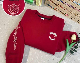 May include: Red sweatshirt with white embroidered details. The front features a set of fangs, while the sleeve has a sword and vines. The back of the sweatshirt says "Back".