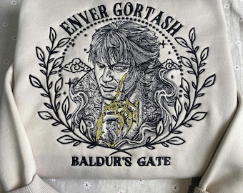 May include: A beige sweatshirt with an embroidered design featuring a character named "Enver Gortash" from the game Baldur's Gate. The design is surrounded by a wreath of leaves and includes a gold-colored detail.