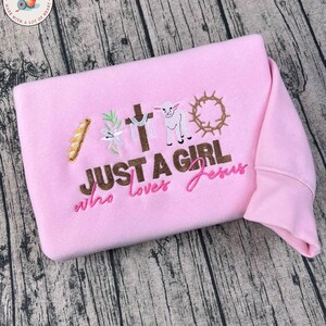 Op de afbeelding: Een lichtroze sweatshirt met geborduurde religieuze symbolen, waaronder een kruis, een lam en een doornenkroon. De tekst "JUST A GIRL who loves Jesus" is ook geborduurd.