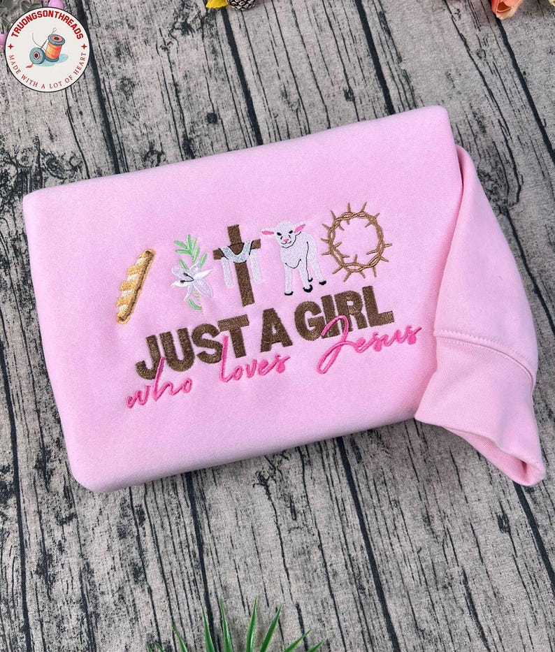 Op de afbeelding: Een lichtroze sweatshirt met geborduurde religieuze symbolen, waaronder een kruis, een lam en een doornenkroon. De tekst "JUST A GIRL who loves Jesus" is ook geborduurd.