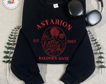 Könnte beinhalten: Schwarzes Sweatshirt mit Rundhalsausschnitt und rotem Stickdesign. Das Design zeigt den Namen "Astarion", den Text "EST. 2023" und "Baldur's Gate" in einer kreisförmigen Grafik. Kopfhörer liegen oben rechts. Die Marke "Truong Son Threads" befindet sich unten.