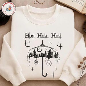 May include: Cream-colored sweatshirt with a black graphic of an umbrella with a forest scene, a howling wolf, and the text "Hoa Hoa Hoa". The design includes stars, crosses, and a heart pendant. The sweatshirt has a crew neck and long sleeves.