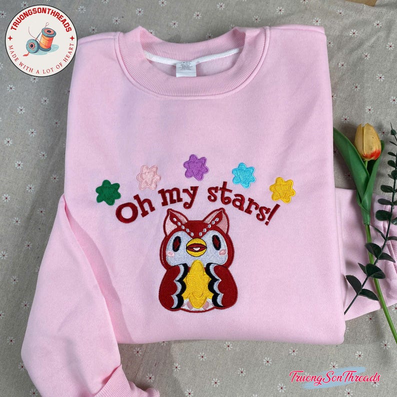 Puede incluir: Una sudadera rosa claro con la frase bordada "Oh my stars!" y un dise&ntilde;o de b&uacute;ho de dibujos animados. El dise&ntilde;o incluye acentos de estrellas de colores. La sudadera tiene cuello redondo y mangas largas.