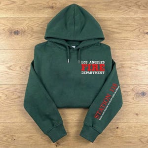 Puede incluir: Una sudadera con capucha de color verde oscuro con cordón ajustable y mangas largas. El pecho muestra el texto en blanco y rojo LOS ANGELES FIRE DEPARTMENT. La manga izquierda presenta el texto en rojo STATION 118 y nombres en blanco.