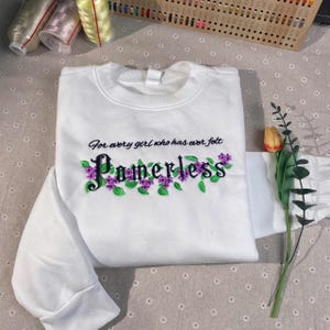 Può includere: Felpa bianca con il testo "For every girl who has ever felt Powerless" ricamato in nero con fiori viola.