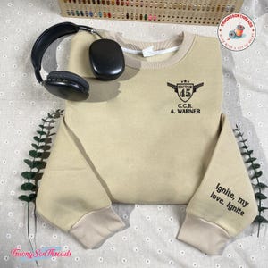 May include: A tan sweatshirt with a crew neck and ribbed cuffs. The sweatshirt features a black graphic on the chest that reads "SECTOR 45 C.C.R. A. WARNER." The right sleeve has the text "Ignite, my love, Ignite." A pair of black headphones rests on the sweatshirt.