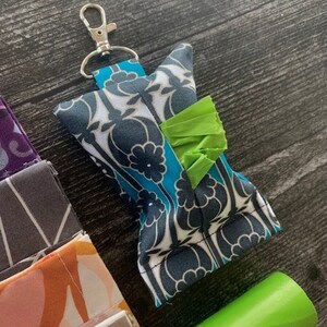 May include: A teal and white patterned fabric dog waste bag dispenser with a green bag attached. The dispenser has a silver clip and is attached to a key ring. A roll of green dog waste bags is also in the image.