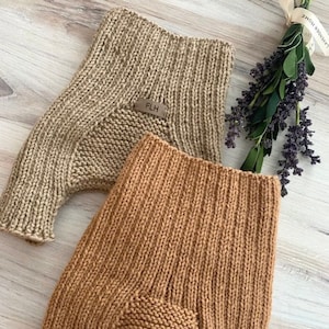 May include: Two pairs of knitted baby pants in tan and light brown. The pants have a ribbed texture and a small leather tag with the initials "FLH". The pants are made of a soft, warm material, perfect for keeping a baby cozy.