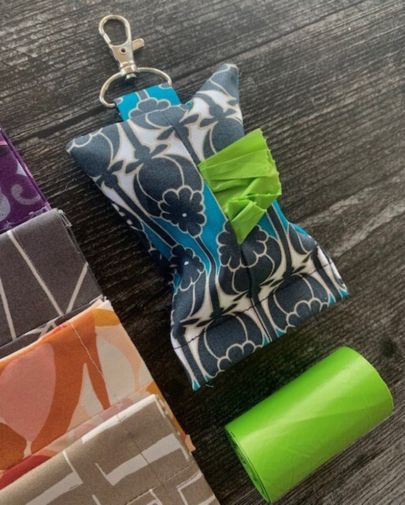 May include: A teal and white patterned fabric dog waste bag dispenser with a green bag attached. The dispenser has a silver clip and is attached to a key ring. A roll of green dog waste bags is also in the image.