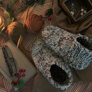May include: Two pairs of knitted grey and brown slippers with a textured pattern. The slippers are on a wooden surface with a book and other items.
