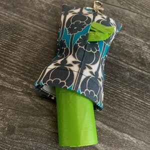 May include: A teal, black, and white patterned fabric dog waste bag dispenser with a green roll of bags inside. The dispenser has a clip for attaching to a leash or belt.