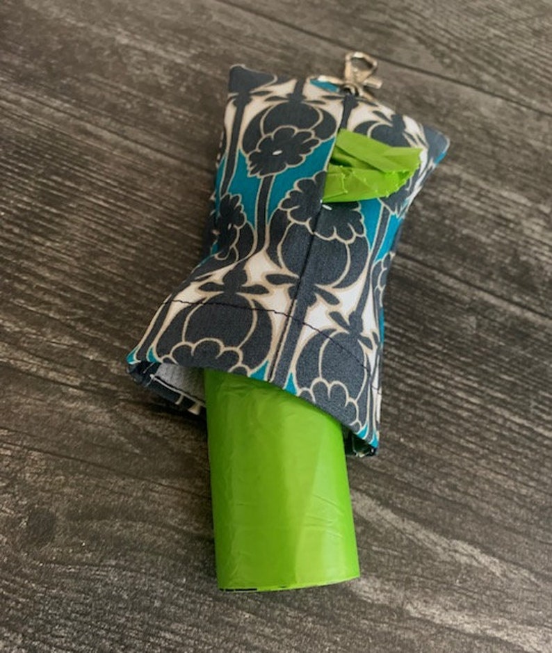May include: A teal, black, and white patterned fabric dog waste bag dispenser with a green roll of bags inside. The dispenser has a clip for attaching to a leash or belt.