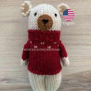 Peut inclure: Un ours en peluche blanc tricot&eacute; portant un pull rouge avec des accents de flocons de neige blancs. Le pull a un col roul&eacute; et l'ours a des yeux en feutre brun et un nez en feutre brun.