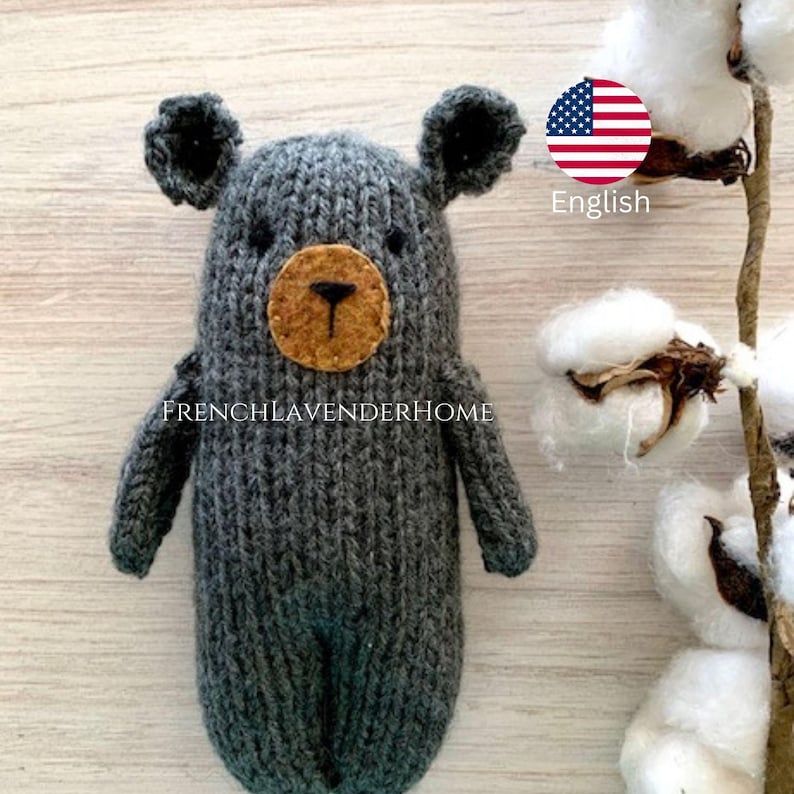 Peut inclure: Un ours en peluche gris tricot&eacute; avec un nez et des yeux en feutre brun. L'ours est debout sur une surface en bois clair avec une plante de coton en arri&egrave;re-plan.