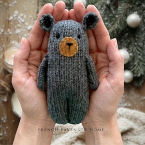 Puede incluir: Un pequeño oso de juguete tejido a mano en color gris oscuro, con un hocico redondo de fieltro color canela, nariz y boca bordadas en negro y dos orejas pequeñas y redondeadas. El oso presenta un patrón de tejido acanalado y texturizado.